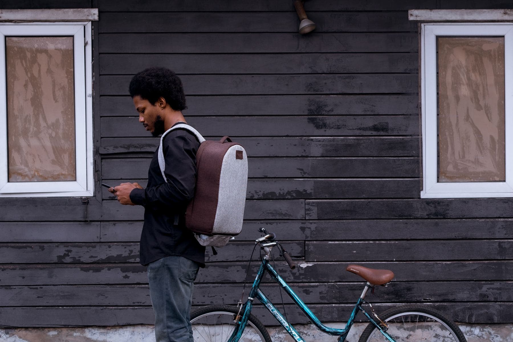 A man with a brown heathered Lightbulb laptop backpack checking his phone beside a bicycle