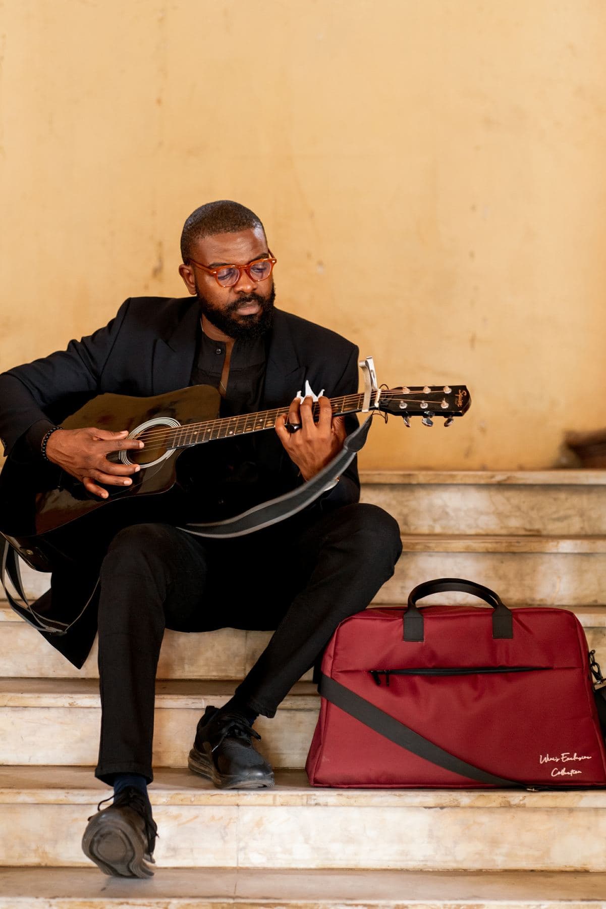 A man playing guitar on warm stone steps beside a crimson Lightbulb weekender bag